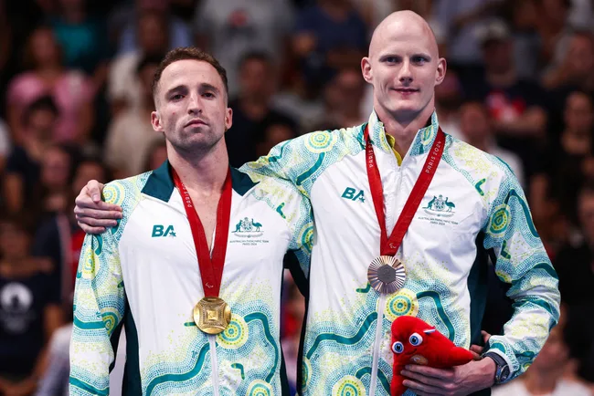 Two athletes in Australian tracksuits with gold and silver medals, standing on a podium, with a plush toy held by one.