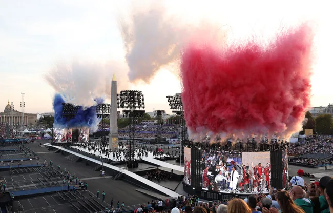 Outdoor ceremony with red, white, and blue smoke, large crowd, stage, and obelisk, likely a public event or celebration.