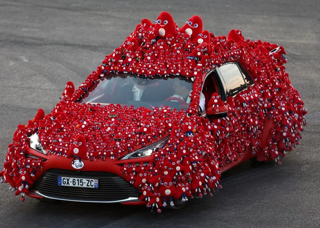 A car covered entirely with red mascots, displaying various expressions, on a paved surface.