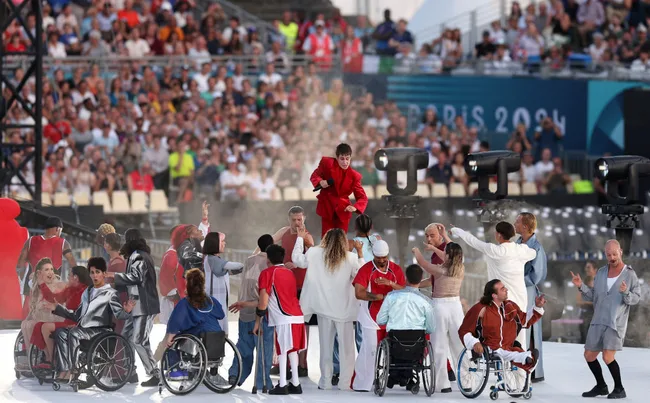 Performers, including wheelchair users, on stage at a sports venue with a Paris 2024 banner, entertaining a large audience.