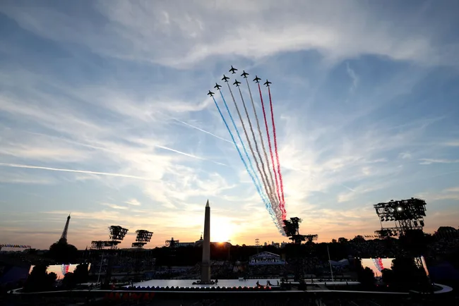 Aerial display with jets trailing blue, white, and red smoke over Paris skyline at sunset, featuring the Eiffel Tower.