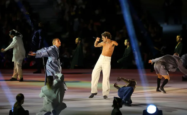 Performer singing shirtless on stage with dancers in robes during a live event.