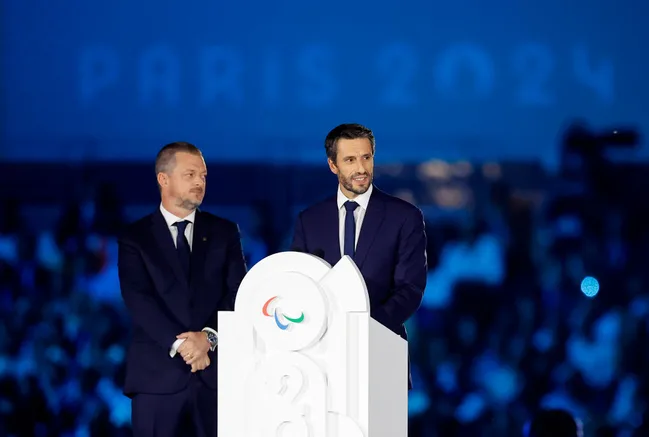 Two men in suits speaking at a podium with the Paris 2024 logo, blurred crowd and "PARIS 2024" text in the background.