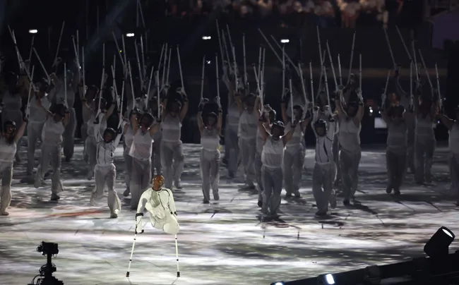 Performer on crutches leads dancers in white outfits holding light sticks, on a lit stage at a major event.