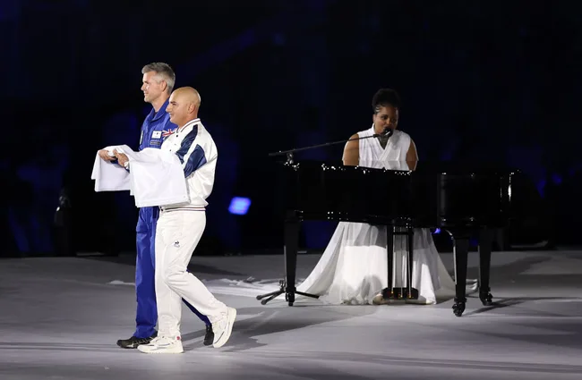 Two individuals in sports attire hold cloths on stage while a woman in white plays piano and sings.