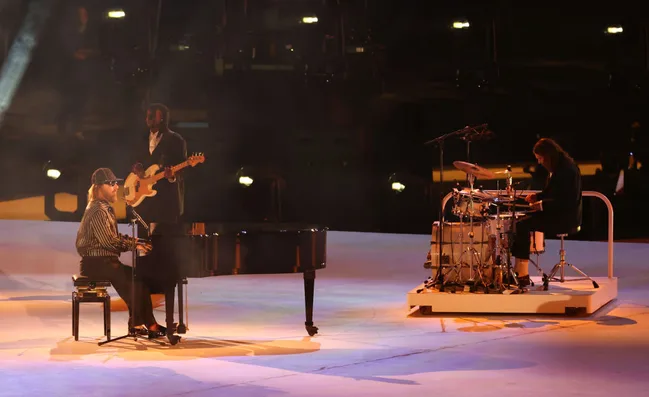 Musicians performing on stage; pianist at a grand piano, guitarist standing, drummer seated at a drum kit.