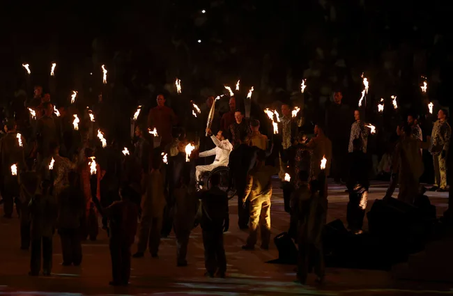 Person in wheelchair holding torch, surrounded by performers with torches at a nighttime event.