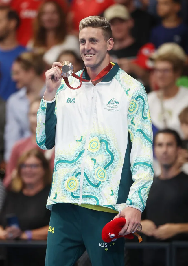 Australian swimmer in a patterned jacket holding a bronze medal with a crowd in the background.