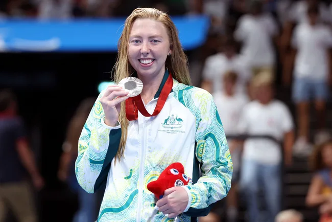 A swimmer wearing an Australian jacket holds a silver medal and a red plush toy, smiling at a swimming event.