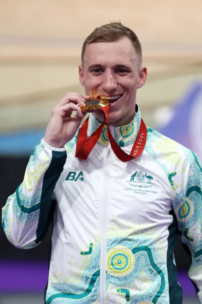 Male athlete in Australian team jacket bites a gold medal, smiling.