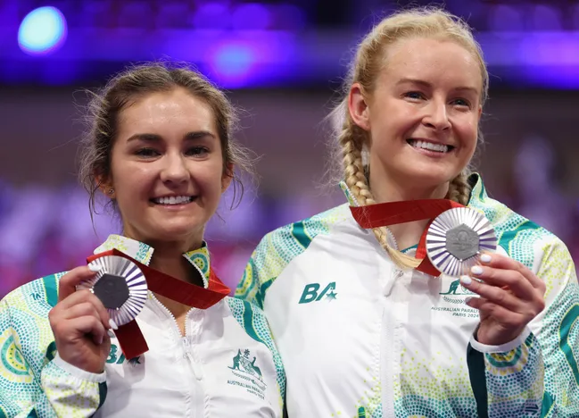 Two female athletes in Australian uniforms smiling, holding silver medals at a sports event.