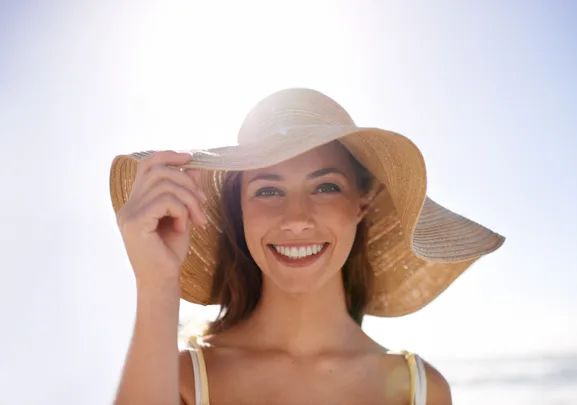 A woman smiling, wearing a large sun hat, with sunlight in the background.