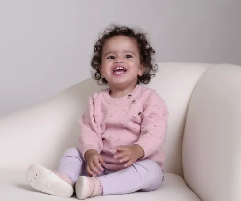 Toddler Tahlia is smiling and wearing pastel pink jumper, sitting on a cream armchair.