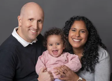 Smiling family portrait of a man, woman, and baby against a gray background.