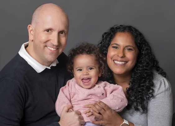 Smiling family portrait of a man, woman, and baby against a gray background.