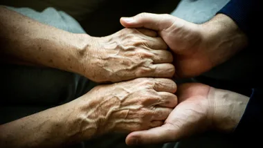 A younger person gently holds an elderly person's hands, symbolizing care and support.