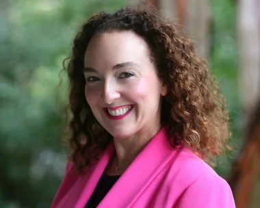 Smiling woman with curly hair wearing a bright pink blazer, standing in front of a lush green background.