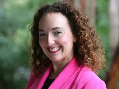 Smiling woman with curly hair wearing a bright pink blazer, standing in front of a lush green background.