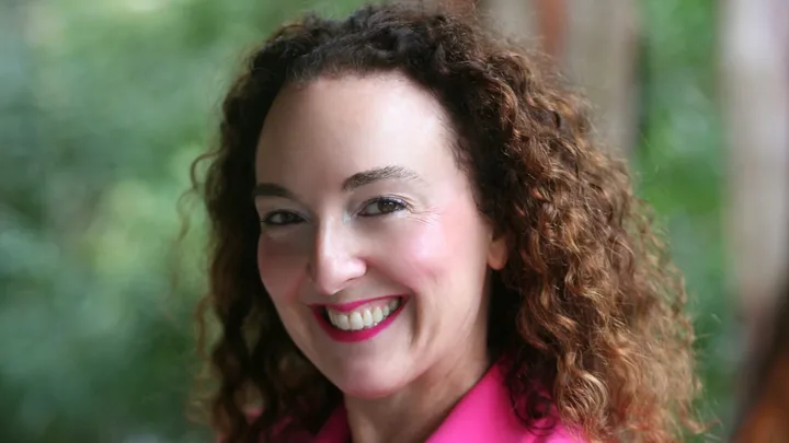 Smiling woman with curly hair wearing a bright pink blazer, standing in front of a lush green background.
