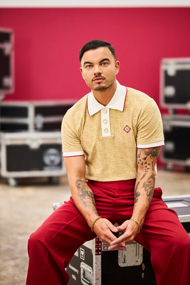 Man with tattoos in a beige polo and red pants, seated on equipment cases against a red background.
