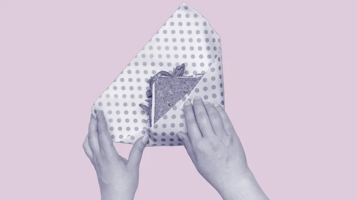 Hands wrapping a sandwich in polka-dot paper against a light pink background.