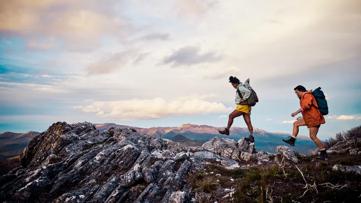 Two hikers with backpacks walk on rocky terrain under a cloudy sky.