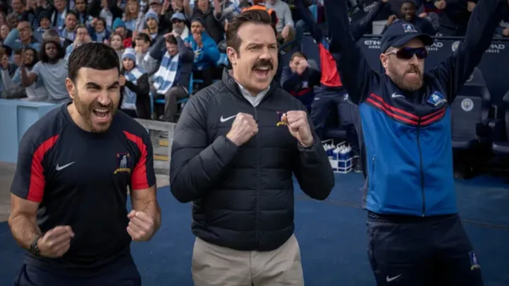 Three men celebrate at a stadium, cheering with excitement, surrounded by a crowd.