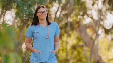 A woman in blue scrubs with a stethoscope stands outdoors, smiling, surrounded by greenery.