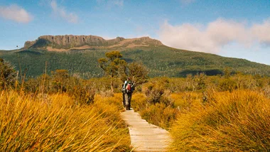 Hiker walking on a wooden trail surrounded by tall grass and mountainous landscape under a clear blue sky.