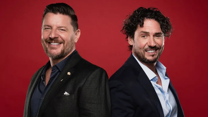 Two smiling men in suits standing back-to-back against a red background.