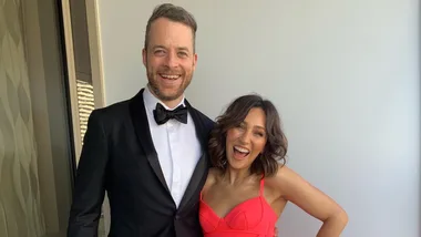 A man in a suit and a woman in a red dress smile broadly, standing side by side against a plain background.