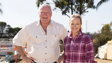 Man and woman smiling at a construction site, wearing casual shirts. Trees and building materials in background.