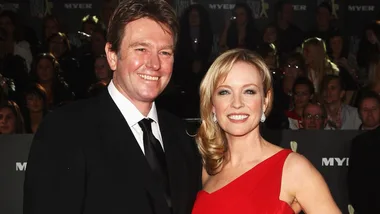 A man in a black suit and a woman in a red dress smiling at an event, with a crowd in the background.