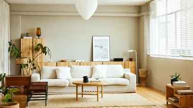 Modern living room with a white sofa, wooden furniture, plants, and large windows with blinds.