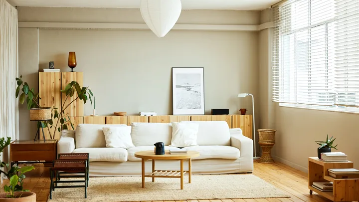 Modern living room with a white sofa, wooden furniture, plants, and large windows with blinds.