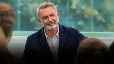 A man with gray hair and a beard, wearing a blue blazer, smiles while interacting with people in a bright setting.