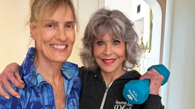 Two women smiling; one holds a blue dumbbell, wearing a black hoodie.