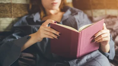 Person in a gray shirt reading a pink book while relaxing on a couch.