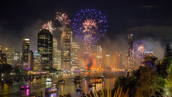 Fireworks explode over a river and city skyline at night, with boats in the water and tall buildings illuminated.