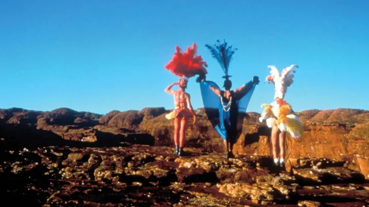 Three people in colorful costumes with feather headdresses stand on a rocky desert landscape under a clear blue sky.