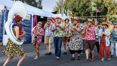 A lively brass band in colorful outfits parades at an outdoor festival with flags and tents in the background.