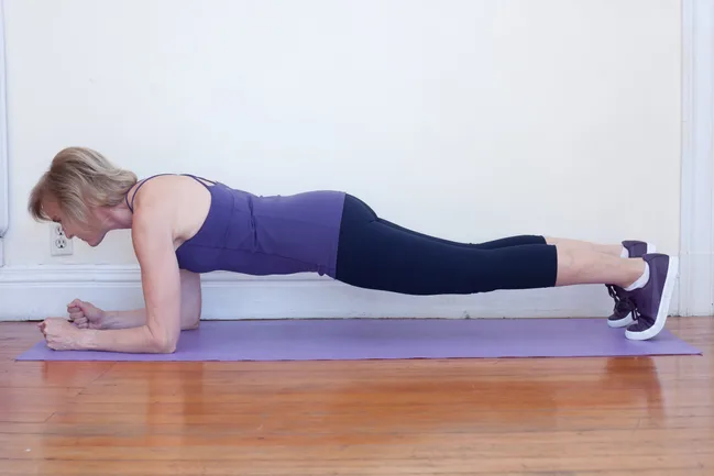 Woman in a purple top and black leggings doing a plank exercise on a yoga mat indoors.