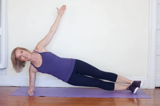 Woman in purple tank top and black leggings doing a side plank on a yoga mat indoors.
