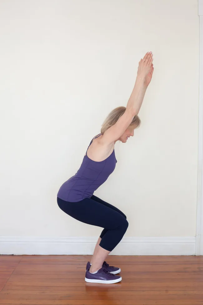 Woman doing yoga chair pose indoors, wearing a purple top and black leggings.