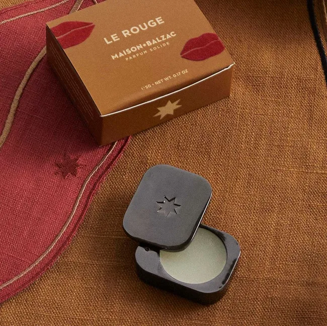 Le Rouge solid perfume by Maison Balzac in a black compact on a textured fabric background with a box.