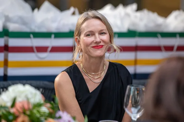 A woman with blonde hair and red lipstick sits outdoors, wearing a black top and gold jewelry, with gift bags in the background.