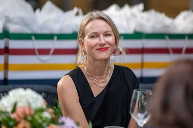 A woman with blonde hair and red lipstick sits outdoors, wearing a black top and gold jewelry, with gift bags in the background.