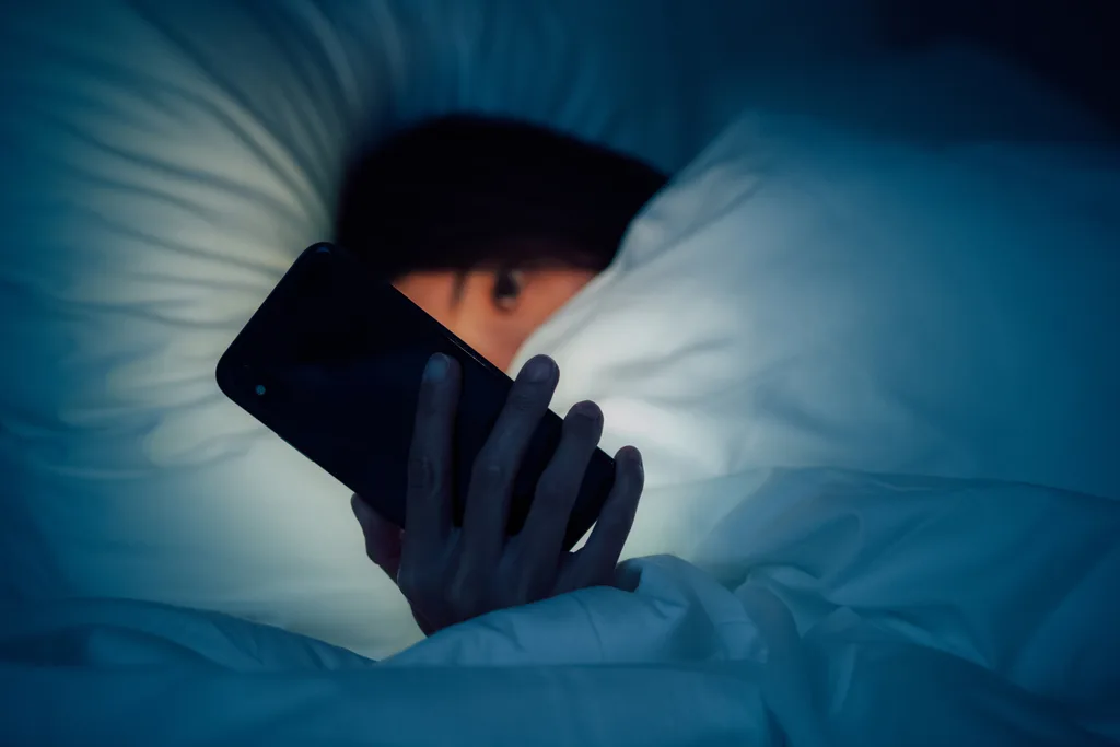 Person under blanket at night, holding a smartphone, face partially covered.