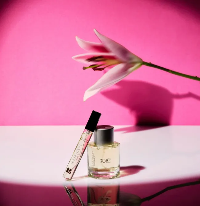 Perfume bottles next to a pink lily on a reflective surface, set against a bright pink background.