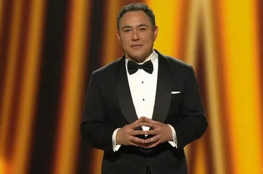 Man in tuxedo standing on stage with golden backdrop.